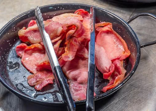 Slices of bacon cooking in a black frying pan with tongs.